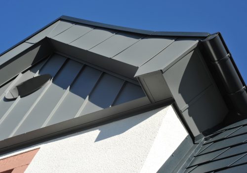 Ecke einer grauen Dachverkleidung auf weisser Fassade mit blauem Himmel und Sonnenschein